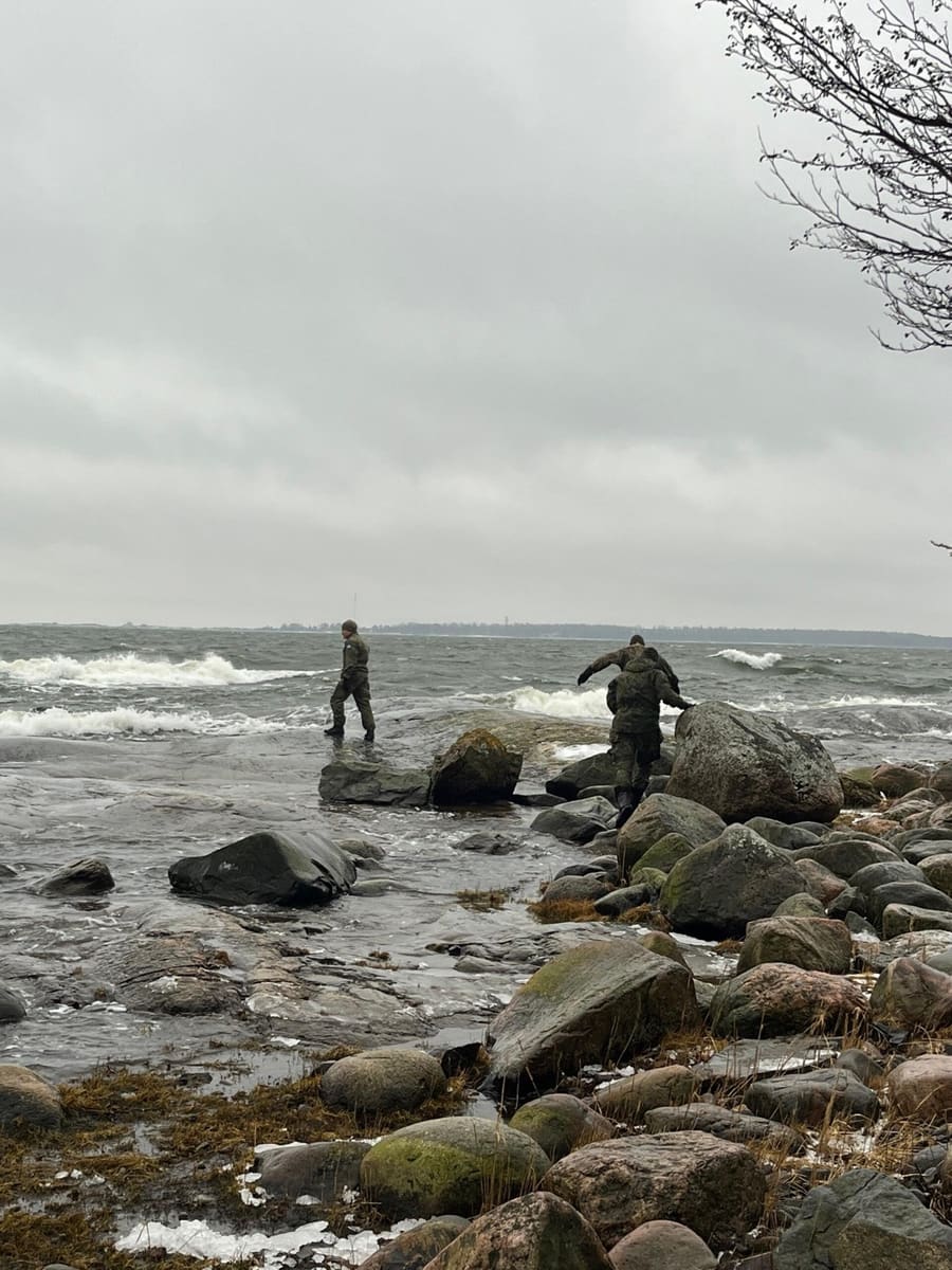 Kaksi henkilöä toimimassa rantakalliolla, aallot taustalla